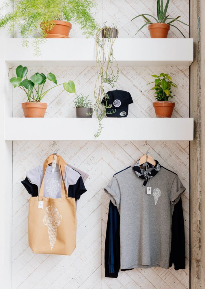White wall with two shelves with various plants and hanging t-shirts and canvas shopping bags for sale in store